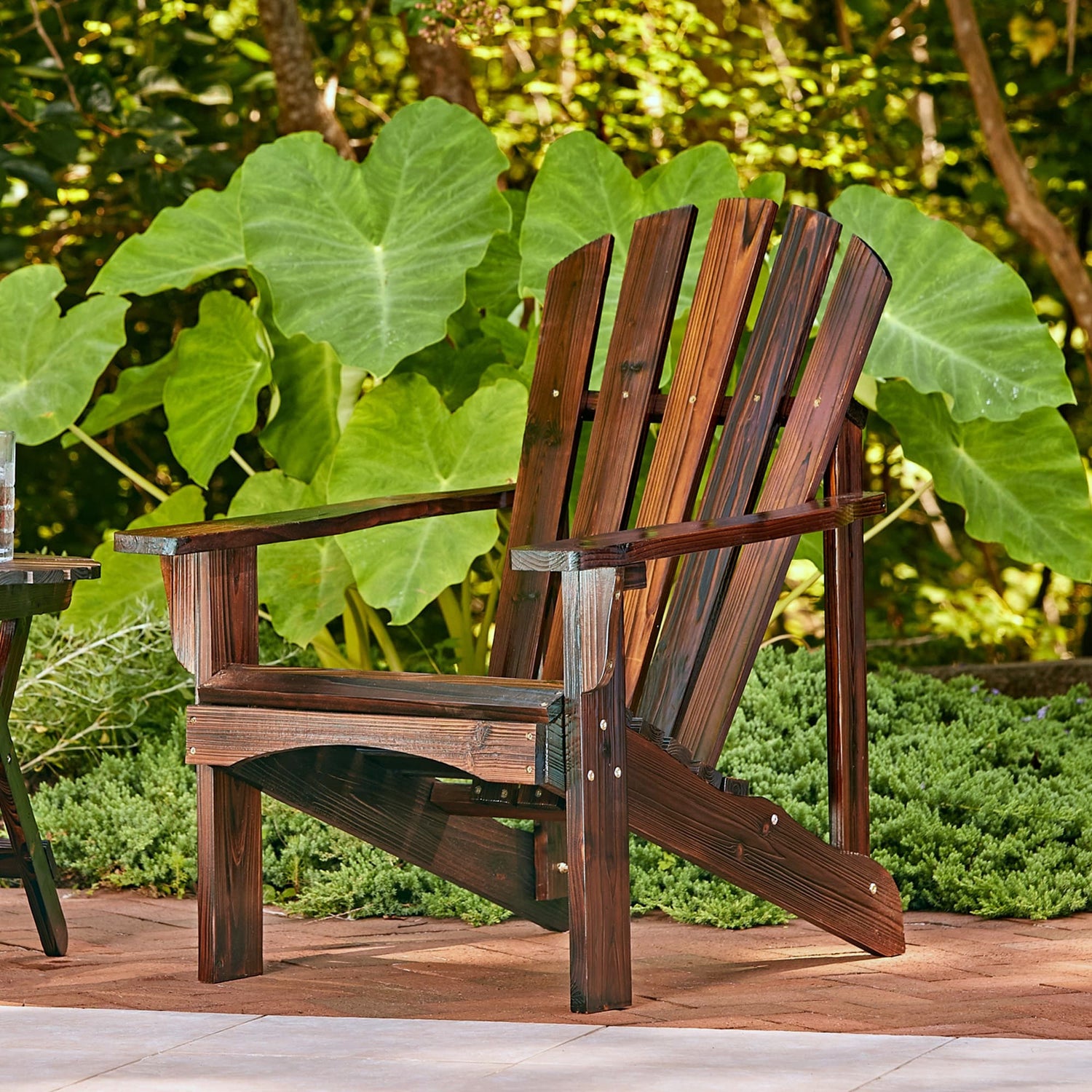 A dark brown wooden Adirondack chair placed on a patio with large green leaves and garden plants in the background. 