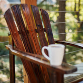 A dark brown wooden Adirondack chair on a porch with a white mug resting on the armrest, set against a blurred outdoor background of trees and a wire railing. 