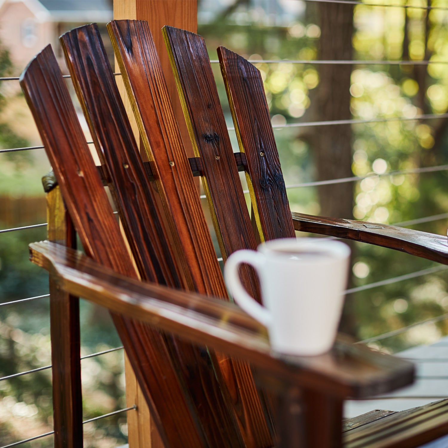 A dark brown wooden Adirondack chair on a porch with a white mug resting on the armrest, set against a blurred outdoor background of trees and a wire railing. 