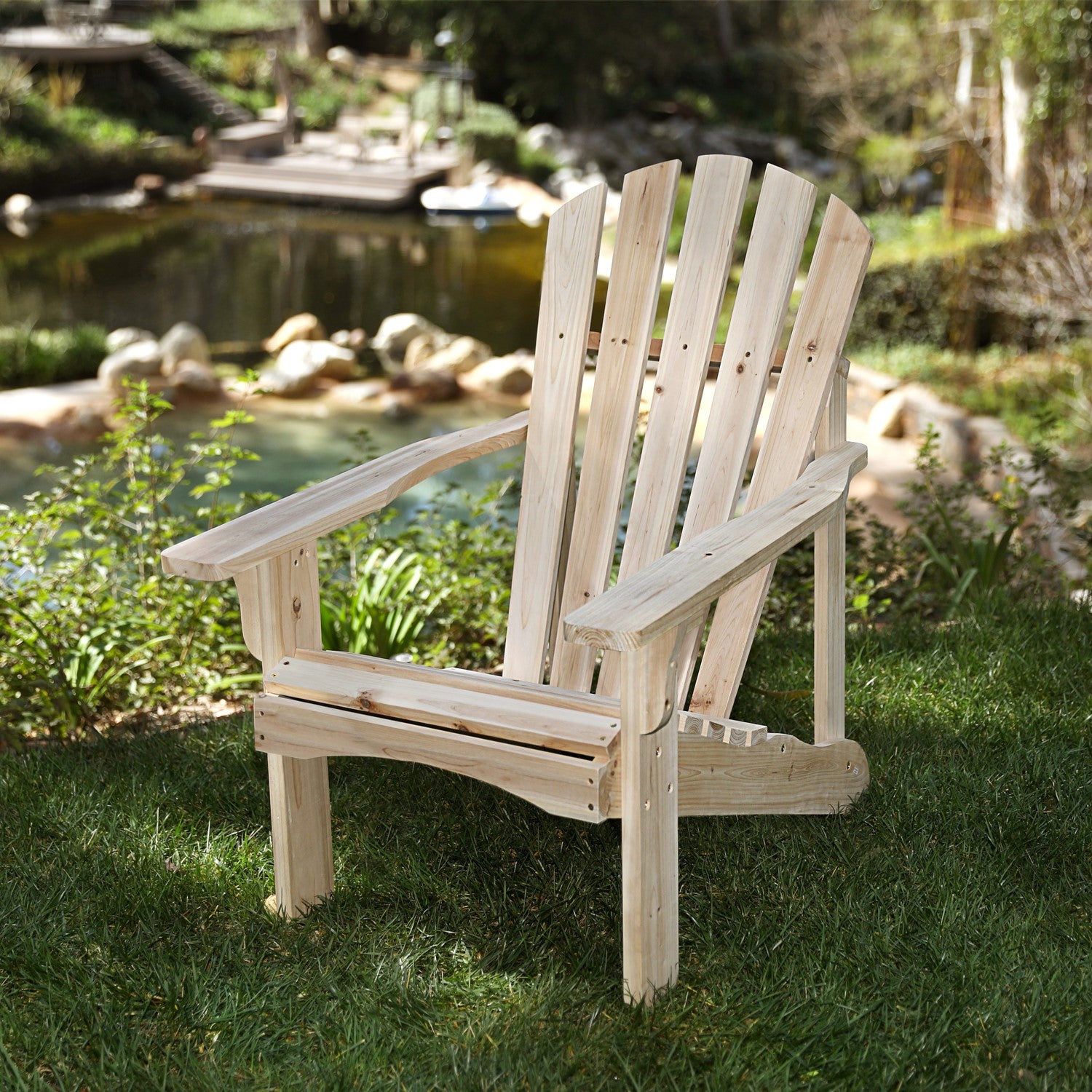 A light natural wood Adirondack chair placed on green grass in a backyard garden setting with a pond and rocks in the background. 