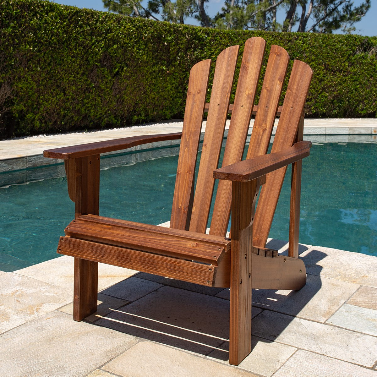 A wooden Adirondack-style outdoor chair with a brown stain, positioned on a stone patio next to a swimming pool with a green hedge in the background. 