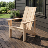 A light natural wood Adirondack chair placed on a dark wooden deck in front of a house with dark siding and a window. 