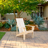 A light natural wood Adirondack-style outdoor chair sits on a stone patio in a landscaped garden with succulents, trees, and a house in the background. 
