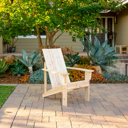 A light natural wood Adirondack-style outdoor chair sits on a stone patio in a landscaped garden with succulents, trees, and a house in the background. 