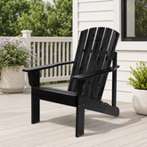A black outdoor Adirondack chair with a slatted back and wide armrests, shown on a light deck beside a white house with potted plants. 