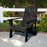 A black outdoor Adirondack-style chair placed on a concrete patio in a garden setting with a white fence, trees, and flowering plants in the background. 