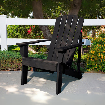 A black outdoor Adirondack-style chair placed on a concrete patio in a garden setting with a white fence, trees, and flowering plants in the background. 