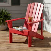 A red Adirondack-style outdoor chair on a wooden deck with a glass of iced tea on the armrest and an open magazine on the seat. 