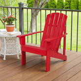 A bright red Adirondack-style outdoor chair sits on a wooden deck beside a white wicker side table with a potted plant, with a railing and green lawn in the background. 