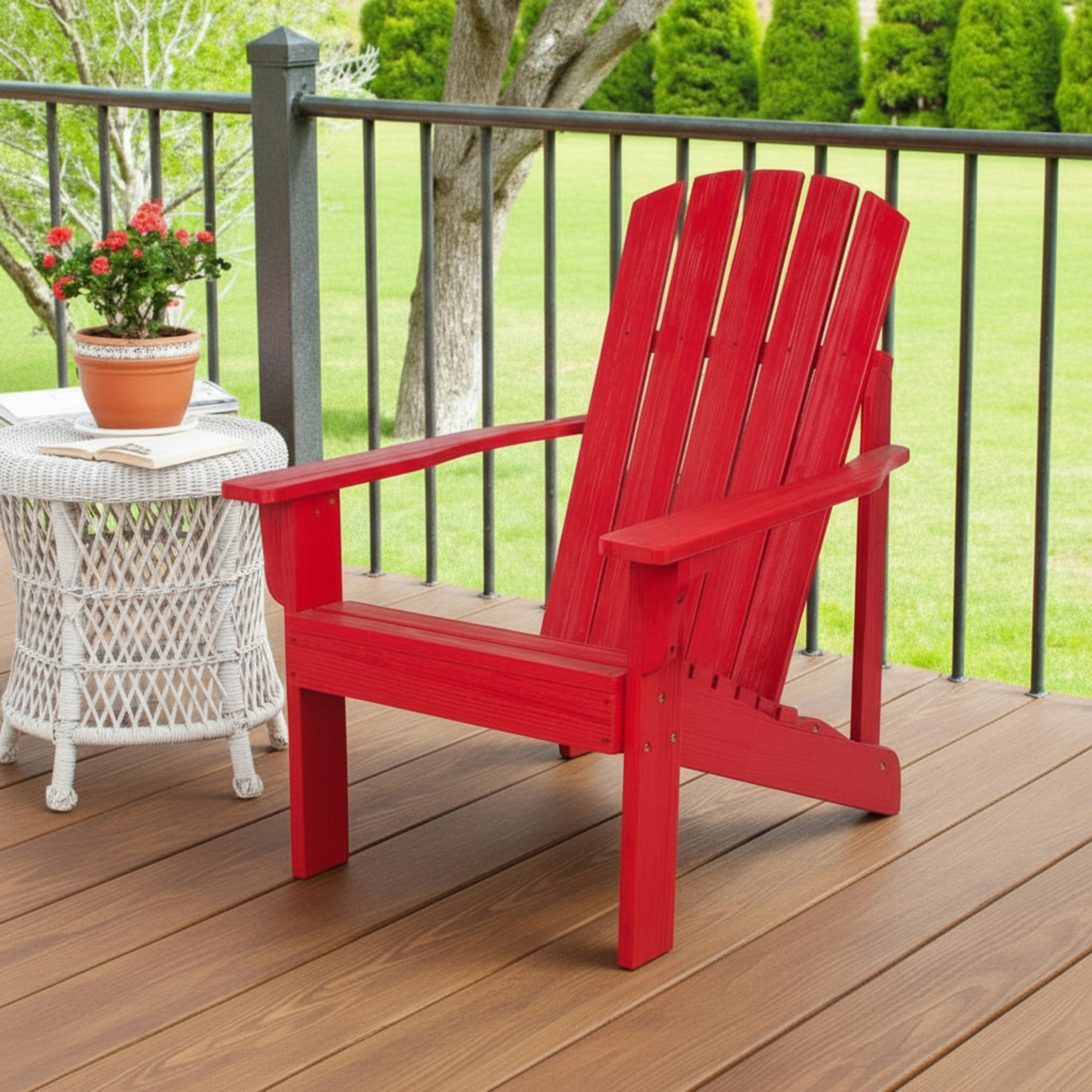 A bright red Adirondack-style outdoor chair sits on a wooden deck beside a white wicker side table with a potted plant, with a railing and green lawn in the background. 