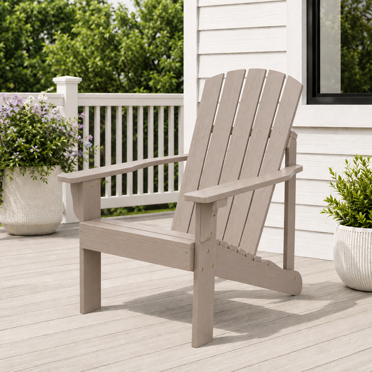 A light grayish-brown outdoor Adirondack chair on a wooden deck next to a white house, with potted plants and a white railing in the background. 