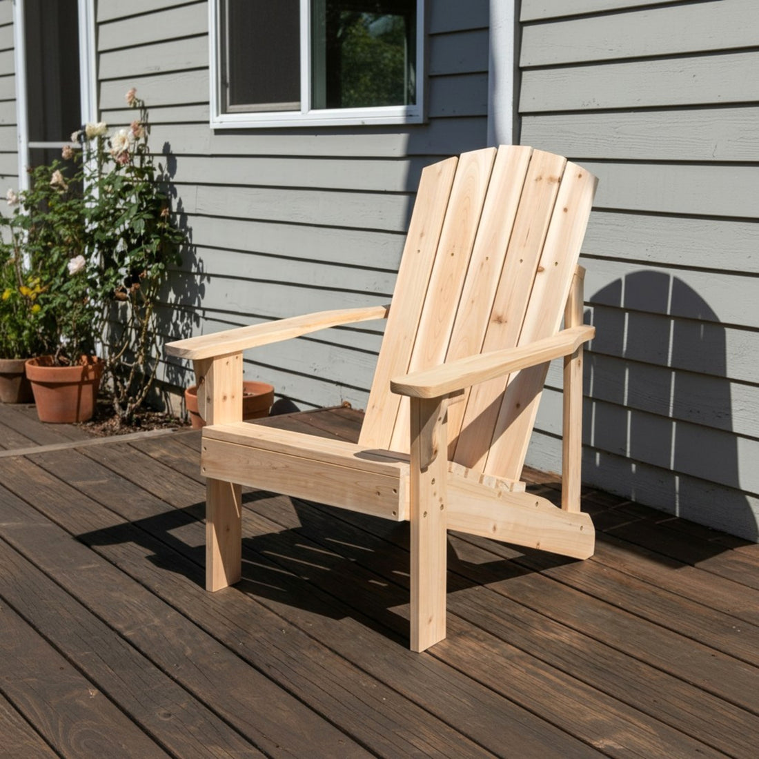 A light natural wood Adirondack chair placed on a dark wooden deck in front of a gray house exterior with potted plants visible on the left. 