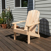 A light natural wood Adirondack chair placed on a dark wooden deck in front of a gray house exterior with potted plants visible on the left. 