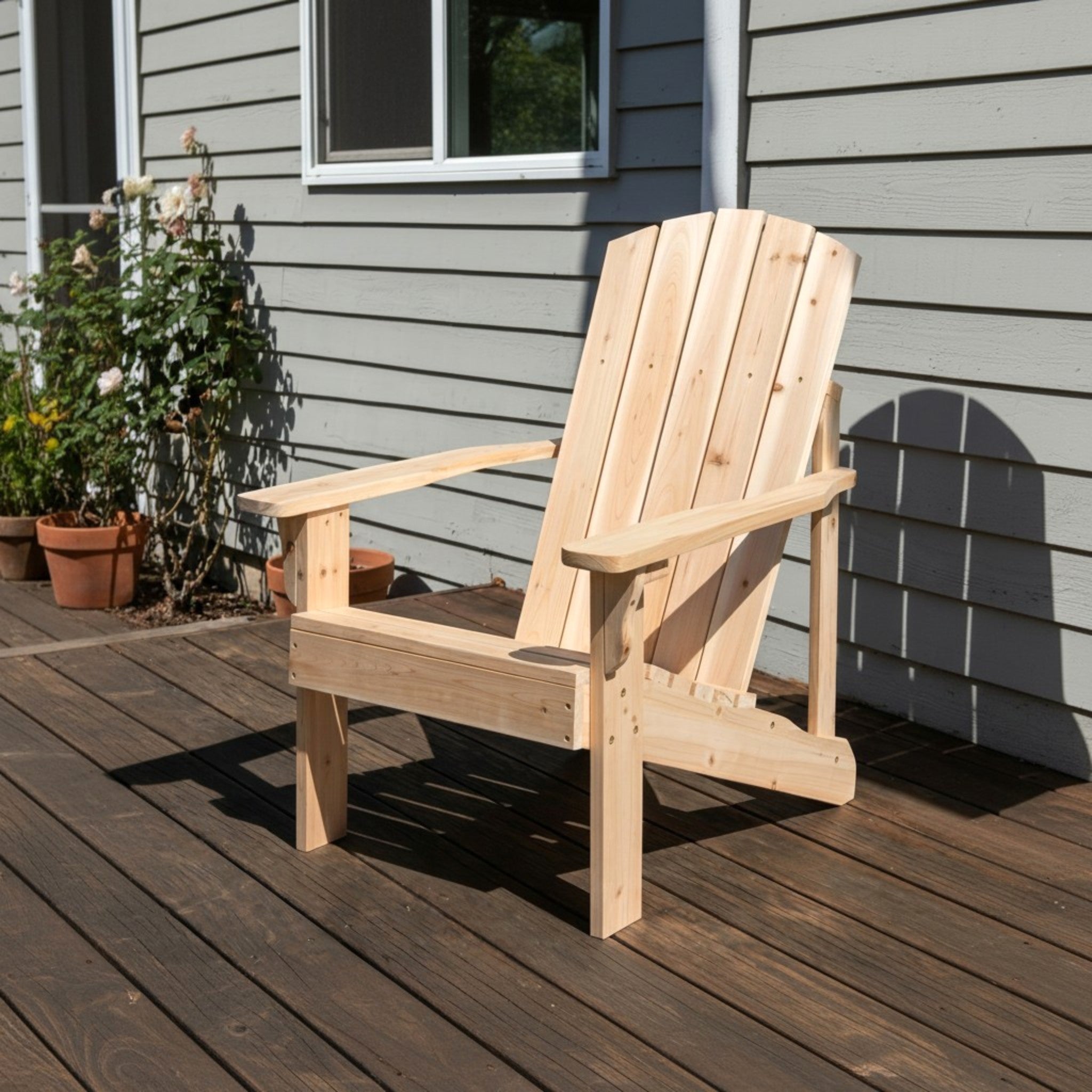 A light natural wood Adirondack chair placed on a dark wooden deck in front of a gray house exterior with potted plants visible on the left. 