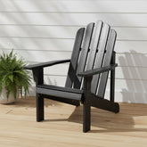 A black outdoor Adirondack chair with a slatted back and wide armrests, shown on a light wood deck against a white siding wall with a potted fern nearby. 