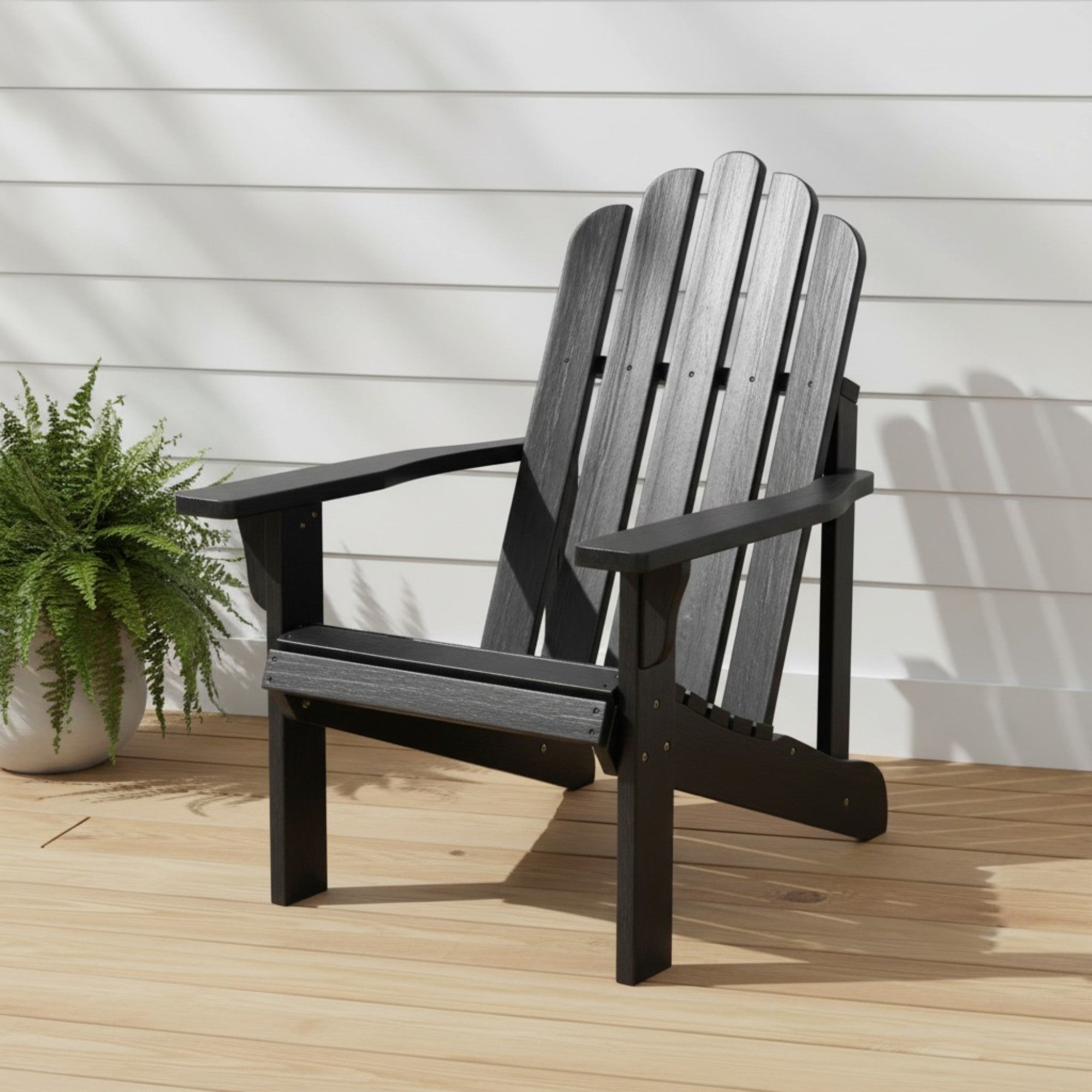 A black outdoor Adirondack chair with a slatted back and wide armrests, shown on a light wood deck against a white siding wall with a potted fern nearby. 