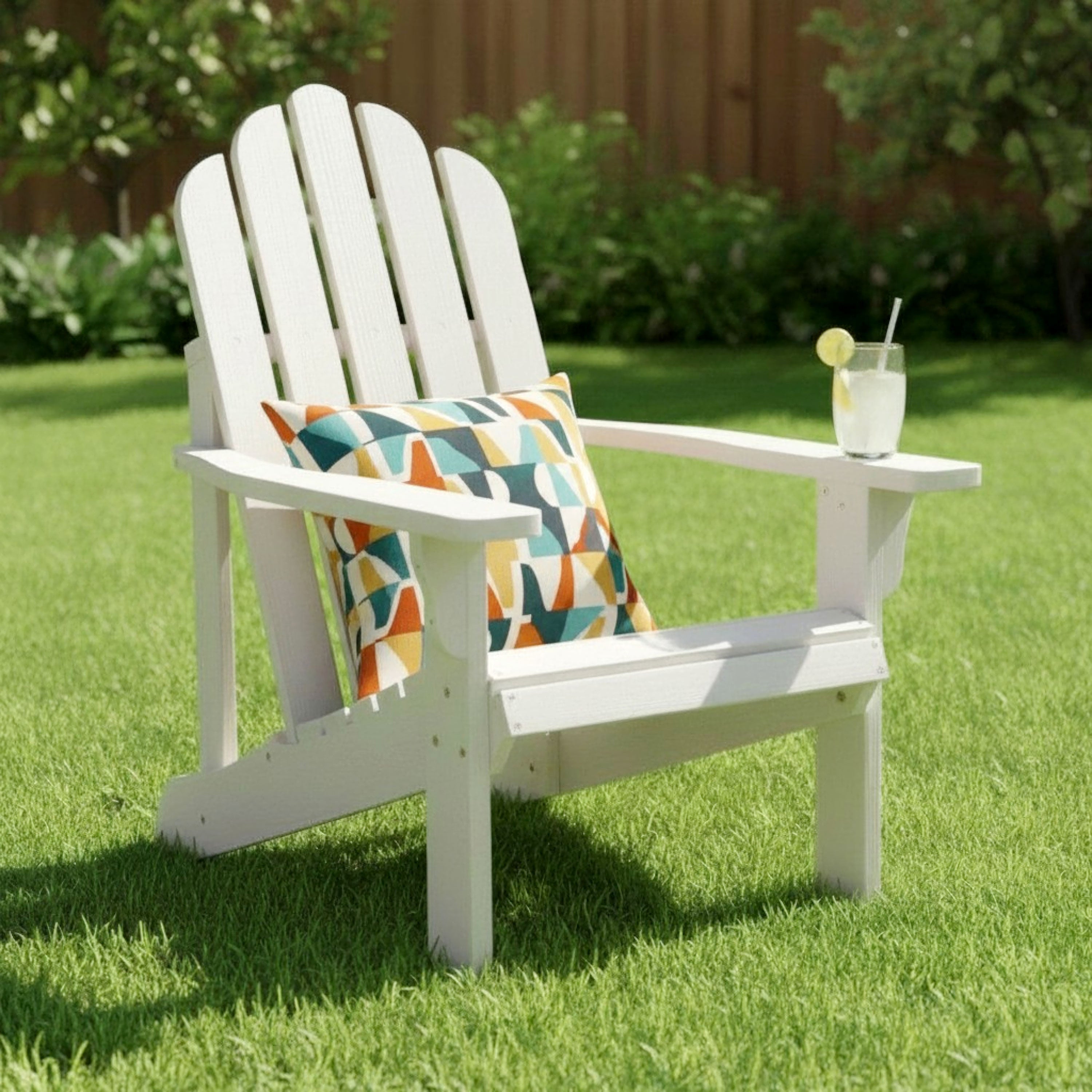 A white outdoor Adirondack chair on a green lawn, styled with a colorful geometric throw pillow and a glass of iced drink on the armrest. 