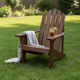 A dark brown wooden Adirondack-style outdoor chair on a green lawn, with a striped throw blanket draped over the arm and a glass of iced tea on the seat. 