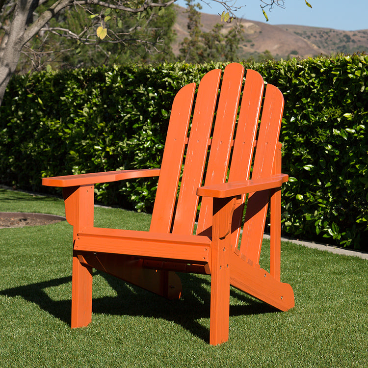 An orange Adirondack-style outdoor chair placed on green grass in front of a dense green hedge, with trees and hills in the background. 