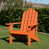 An orange Adirondack-style outdoor chair placed on green grass in front of a dense green hedge, with trees and hills in the background. 