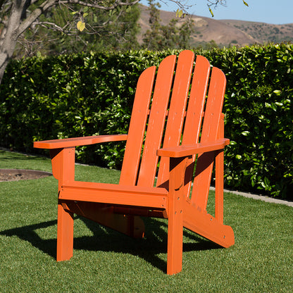 An orange Adirondack-style outdoor chair placed on green grass in front of a dense green hedge, with trees and hills in the background. 