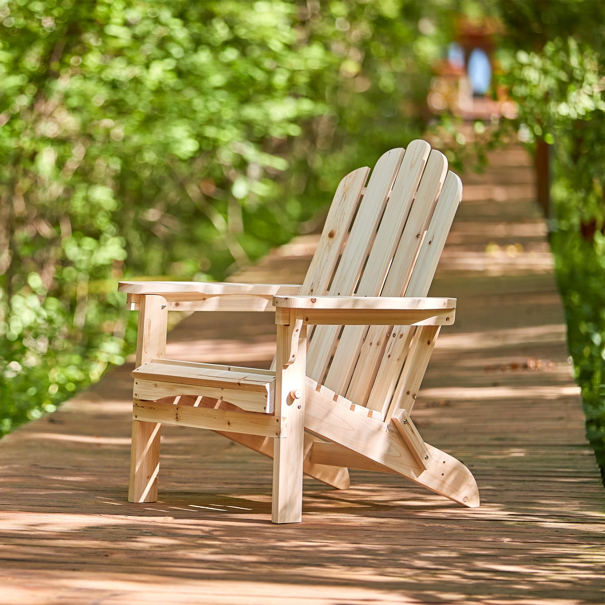 A light natural wood Adirondack chair placed on a wooden boardwalk surrounded by lush green trees and foliage. 