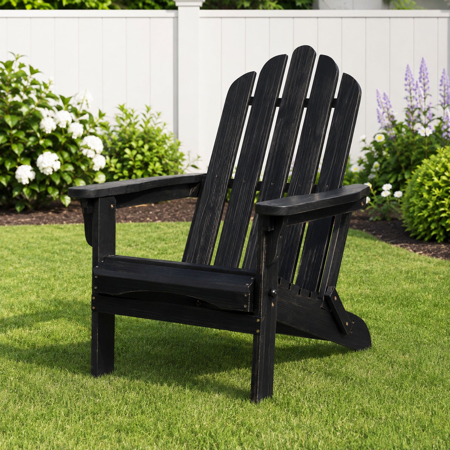 A black wooden Adirondack chair placed on a green lawn in front of a white fence with garden plants. 