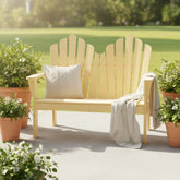 A light pastel yellow wood Adirondack-style outdoor bench with a slatted back, shown on a patio with potted plants and a throw blanket. A cream pillow rests on the bench.
