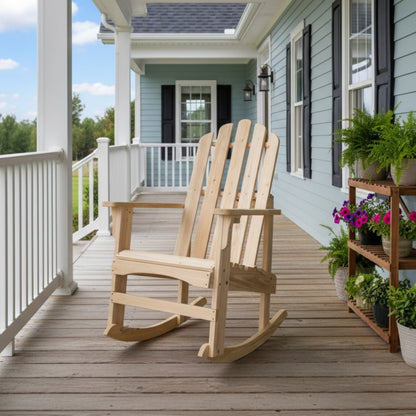 A natural light wood Adirondack-style rocking chair sits on a wooden front porch of a light blue house with white railings and potted plants. 