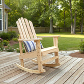 A light natural wood Adirondack-style rocking chair with a blue and white striped throw pillow, placed on a wooden deck in a backyard setting with trees and grass in the background. 