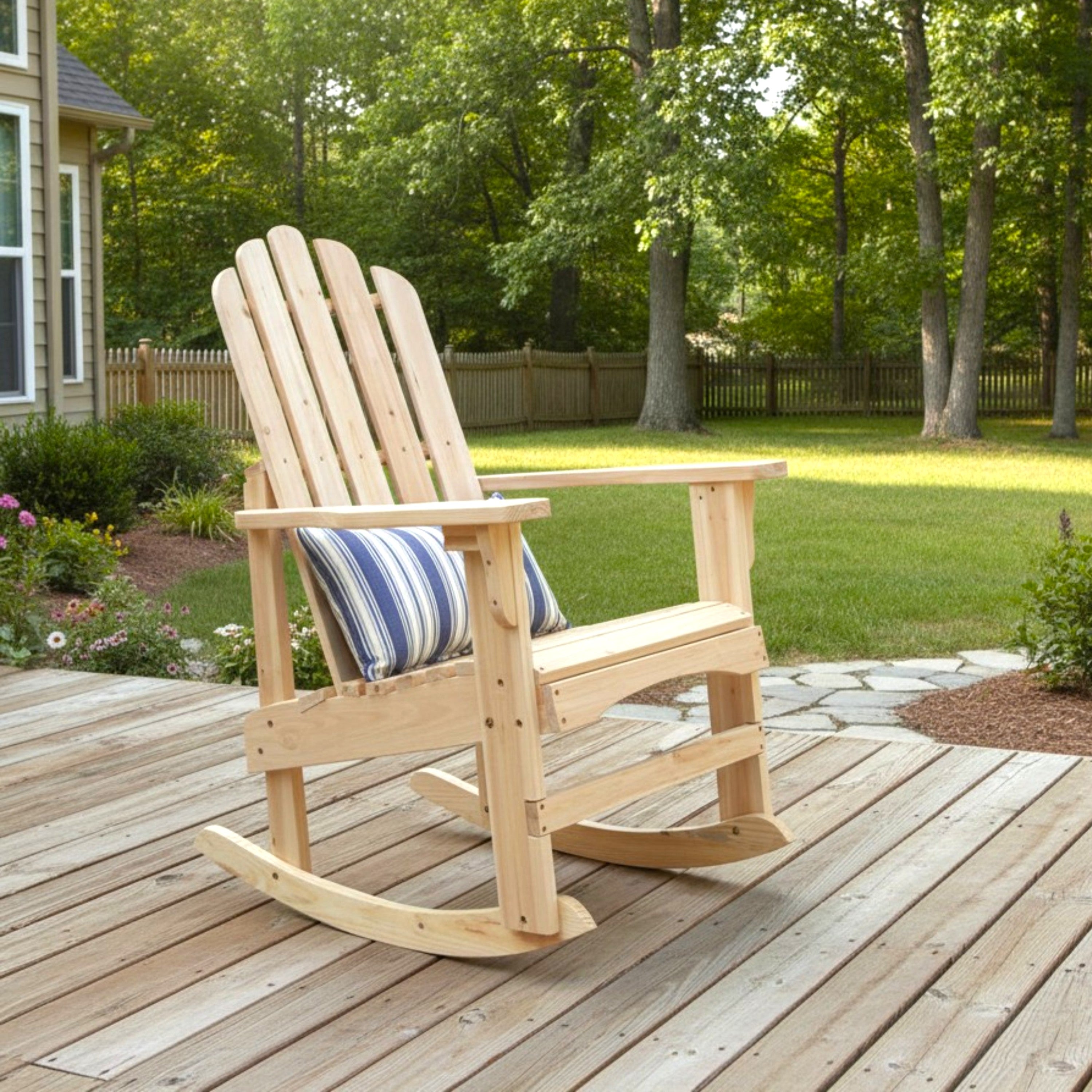 A light natural wood Adirondack-style rocking chair with a blue and white striped throw pillow, placed on a wooden deck in a backyard setting with trees and grass in the background. 
