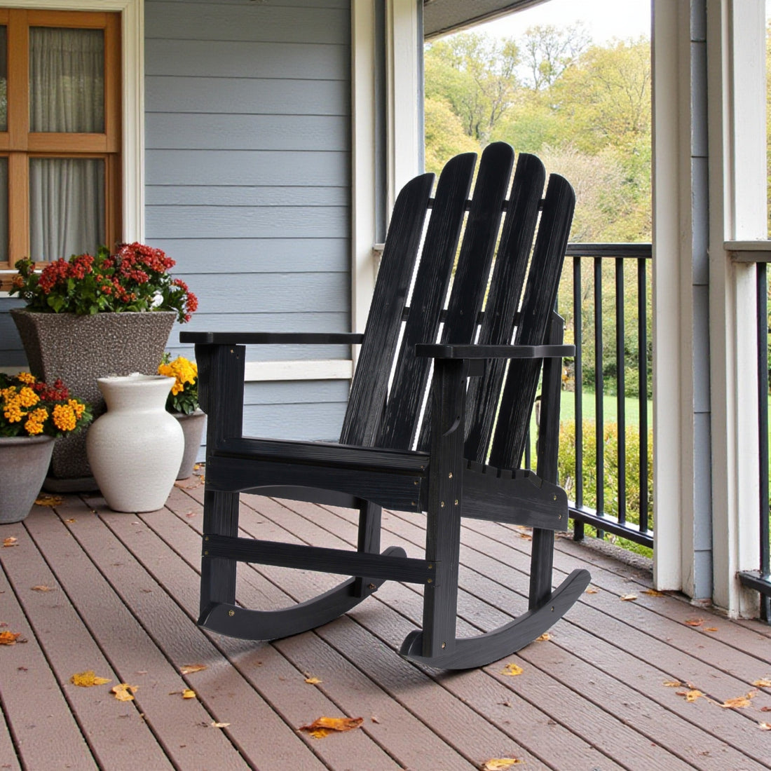 A black Adirondack-style rocking chair sits on a brown wooden porch deck, with potted flowers and a gray house exterior in the background. 