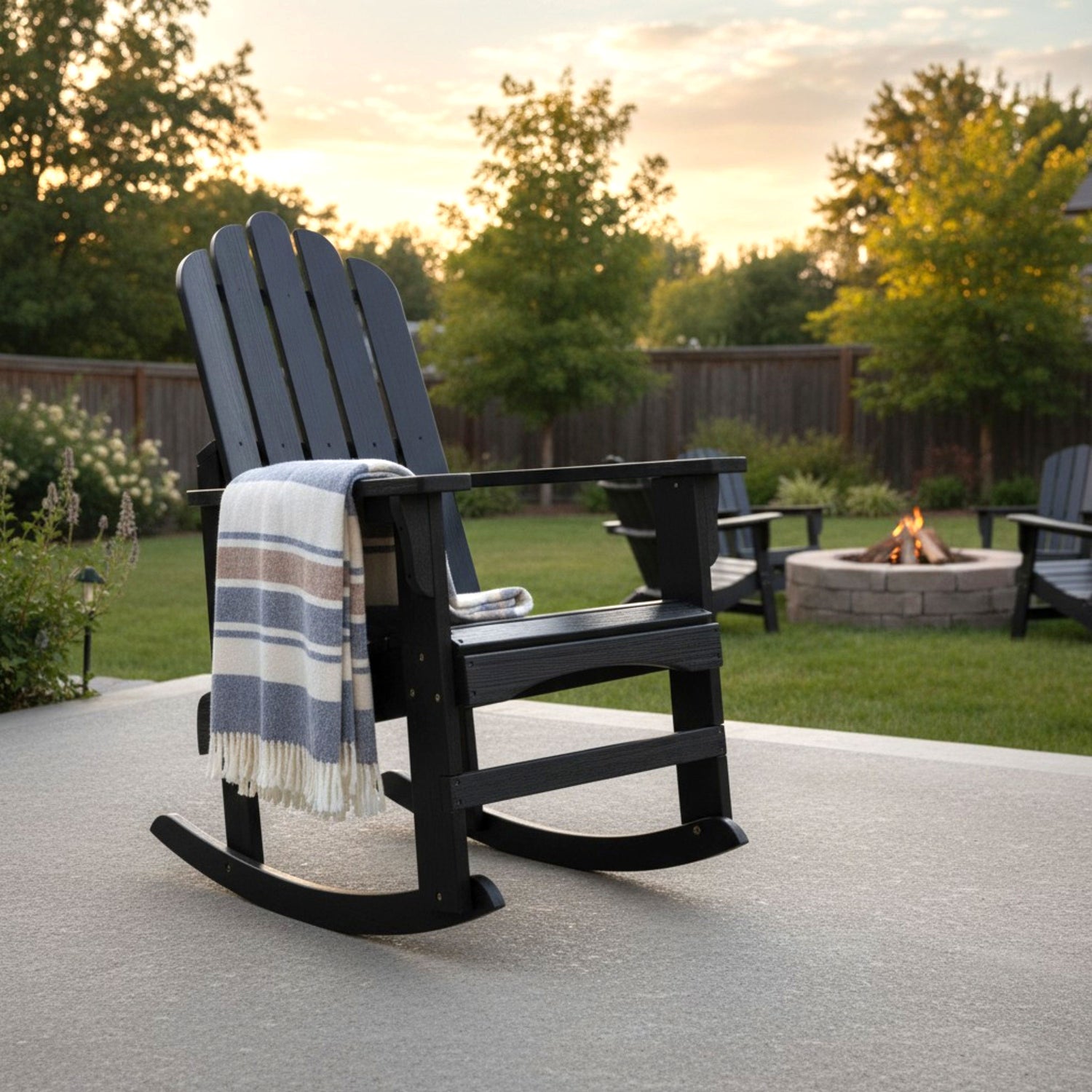 A black Adirondack-style rocking chair on a concrete patio in a backyard setting, with a blue, white, and beige striped throw blanket draped over the arm. Trees, a fire pit, and other patio chairs are visible in the background at sunset. 