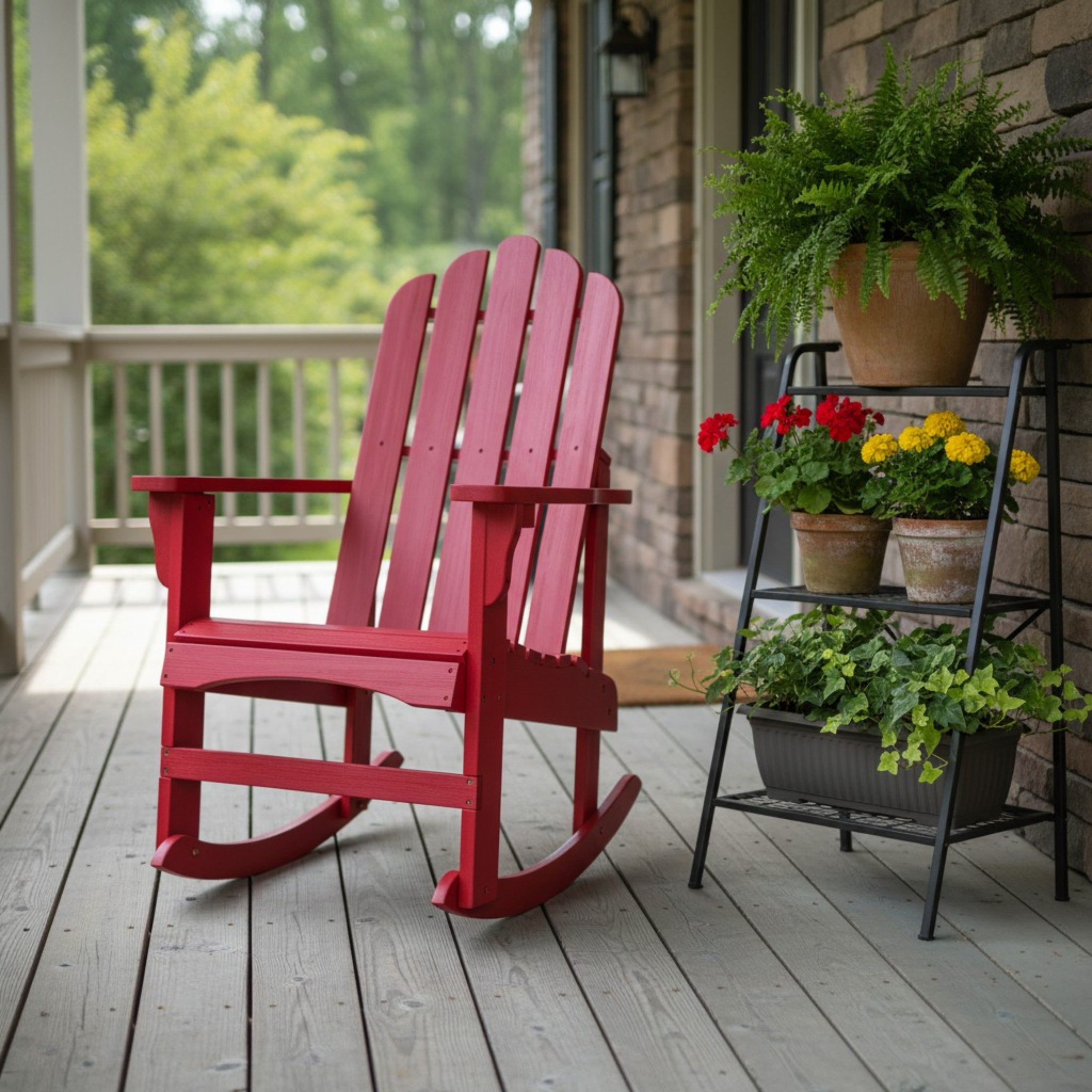 A red wooden Adirondack-style rocking chair sits on a gray deck beside a black metal plant stand holding potted greenery and flowers, with a porch and brick wall in the background. 