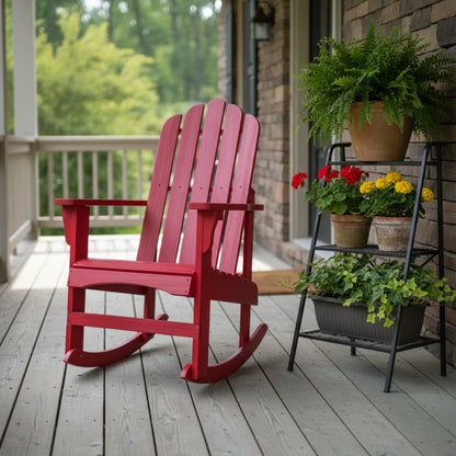 A red wooden Adirondack-style rocking chair sits on a gray deck beside a black metal plant stand holding potted greenery and flowers, with a porch and brick wall in the background. 