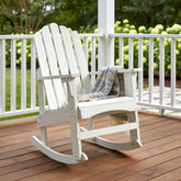A white wooden Adirondack-style rocking chair on a brown deck with a gray patterned throw blanket draped over the seat, set on a porch with greenery in the background. 