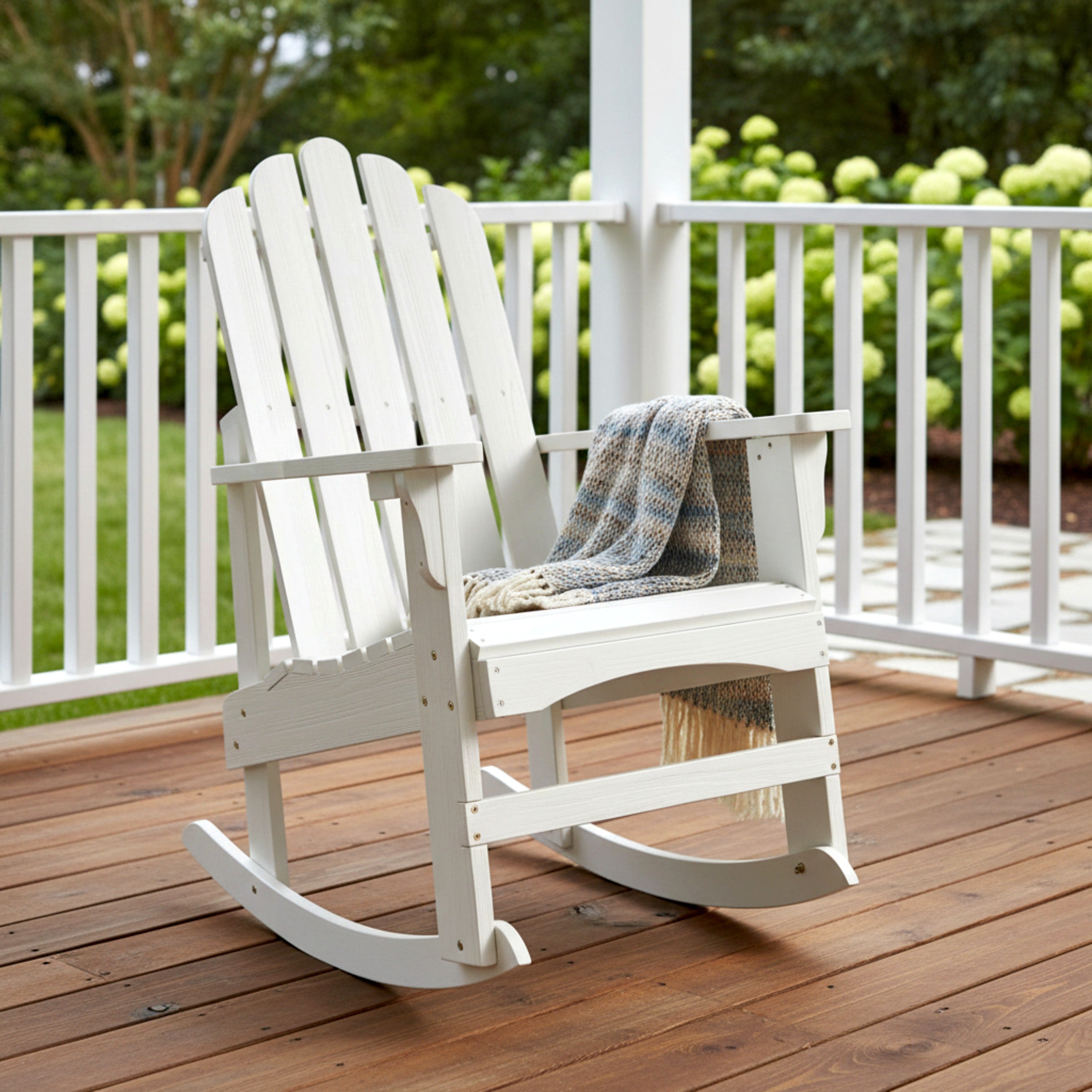 A white wooden Adirondack-style rocking chair on a brown deck with a gray patterned throw blanket draped over the seat, set on a porch with greenery in the background. 