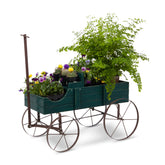 A dark green wooden garden wagon planter with rustic metal wheels and a tall handle, filled with green foliage and colorful flowers, shown on a white background. 