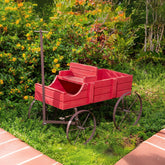 A red wooden garden wagon decoration with dark metal wheels and a vertical post, placed on a grassy lawn in a garden setting with flowering bushes. 