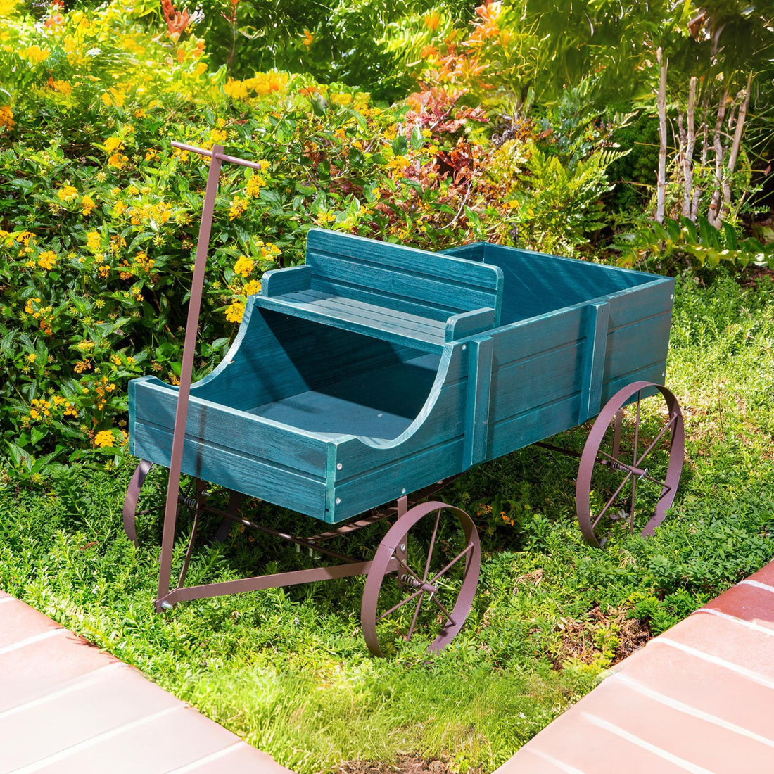 A rustic green wooden garden wagon with dark metal wheels and a handle, placed on a green lawn in a garden setting with yellow flowers and a brick path. 