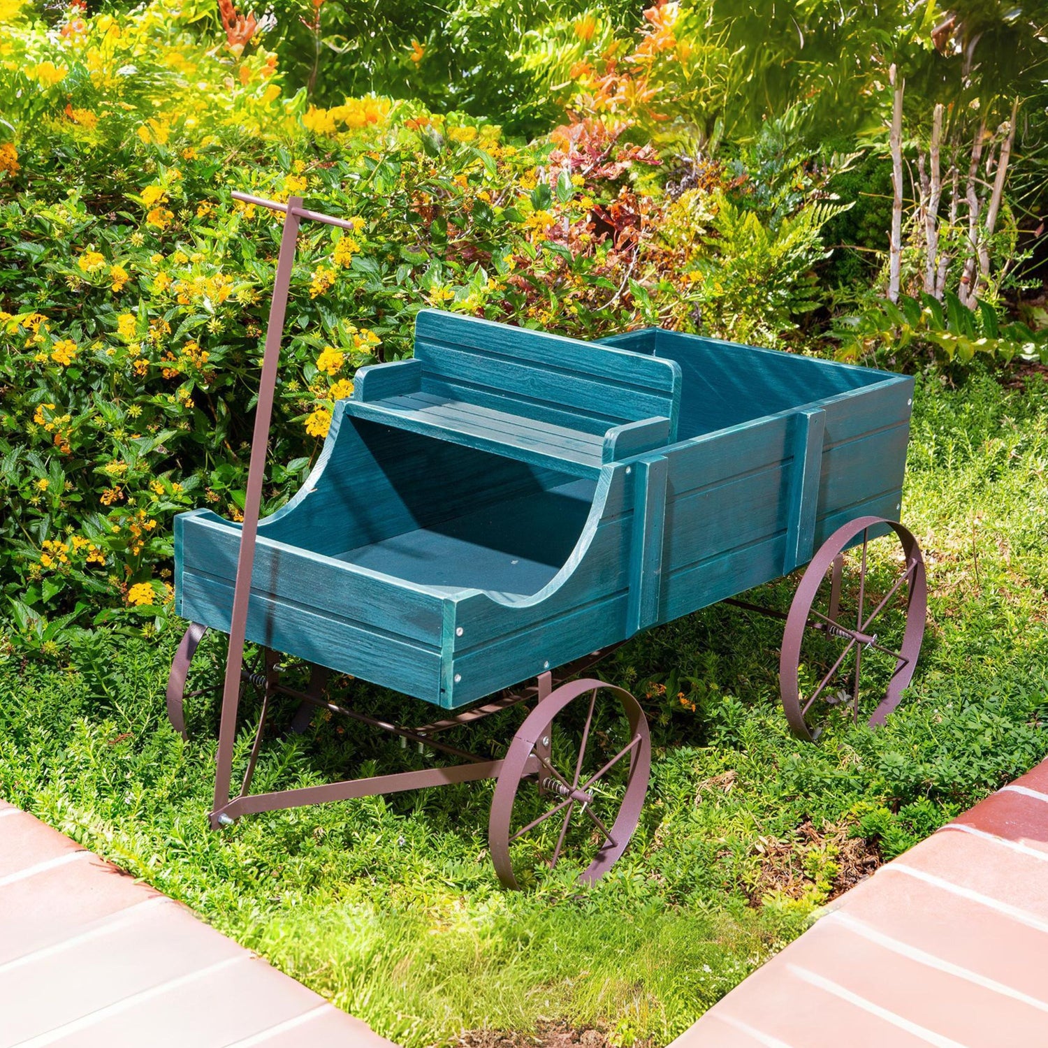 A rustic green wooden garden wagon with dark metal wheels and a handle, placed on a green lawn in a garden setting with yellow flowers and a brick path. 