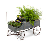 A rustic gray wooden wagon planter with dark metal wheels and a vertical hitch arm, filled with green plants and small potted flowers, shown on a white background. 