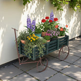 A dark green wooden slat planter cart with a rusted brown metal frame and wheels, filled with various colorful potted flowers and green trailing plants, set on a paved patio against a beige wall with leafy shadows. 