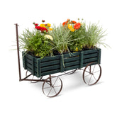 A dark green wooden slat planter wagon with metal wheels and a handle, filled with green ornamental grasses and colorful flowers, photographed on a white background. 