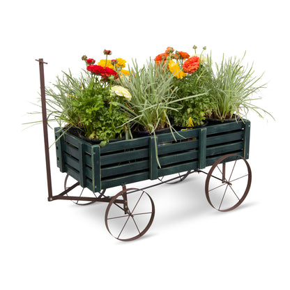A dark green wooden slat planter wagon with metal wheels and a handle, filled with green ornamental grasses and colorful flowers, photographed on a white background. 