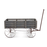 A rustic wooden wagon planter with a weathered gray-brown slatted crate body and dark metal wheels with white-walled tires, shown on a plain white background. 