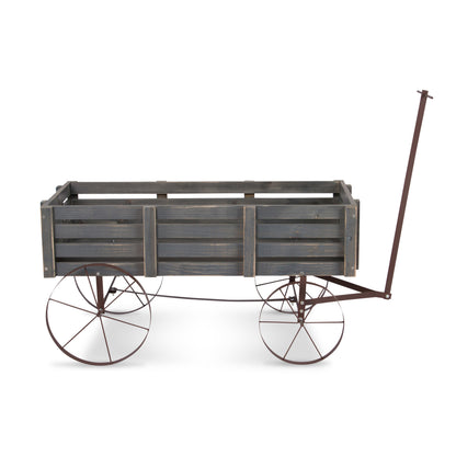 A rustic wooden wagon planter with a weathered gray-brown slatted crate body and dark metal wheels with white-walled tires, shown on a plain white background. 