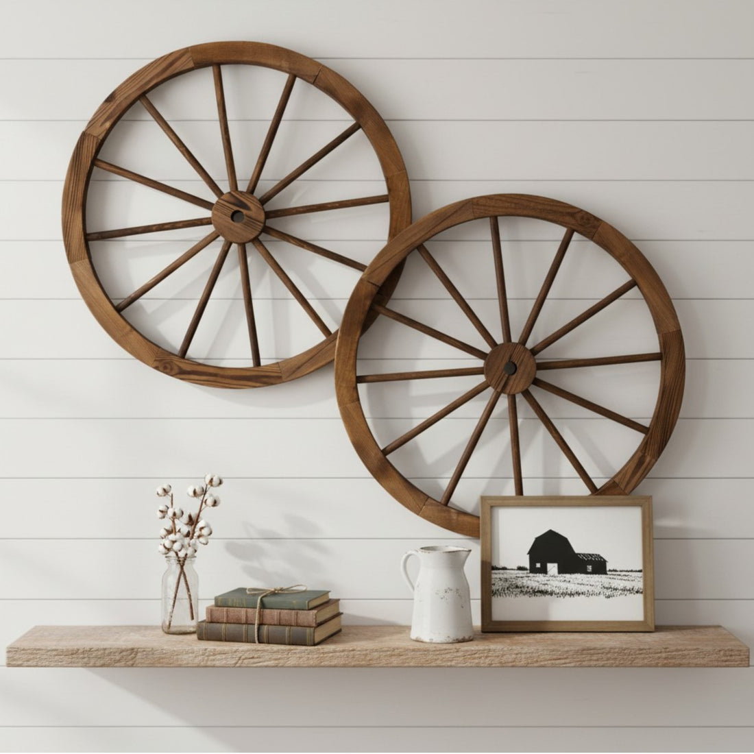 Two large rustic wooden wagon wheel wall decor pieces mounted on a white shiplap wall above a wooden shelf with decorative books, a small vase, and a framed black-and-white barn print. 