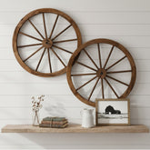 Two large rustic wooden wagon wheel wall decor pieces mounted on a white shiplap wall above a wooden shelf with decorative books, a small vase, and a framed black-and-white barn print. 