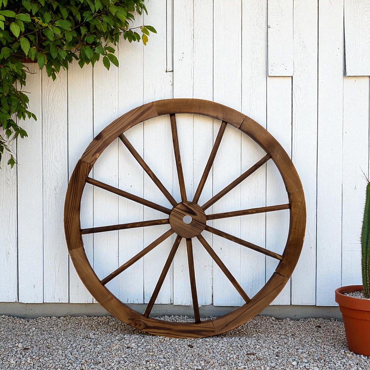 A large rustic wooden wagon wheel wall decor piece leaning against a white wood paneled wall on a gravel surface, with green foliage and a potted cactus nearby. 